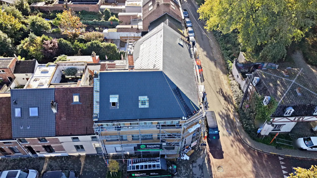 Dakrenovatie in Asse van boven: het nieuwe donkere dak van een hoekhuis, met stelling, spandoek en bestelwagen van Konverto